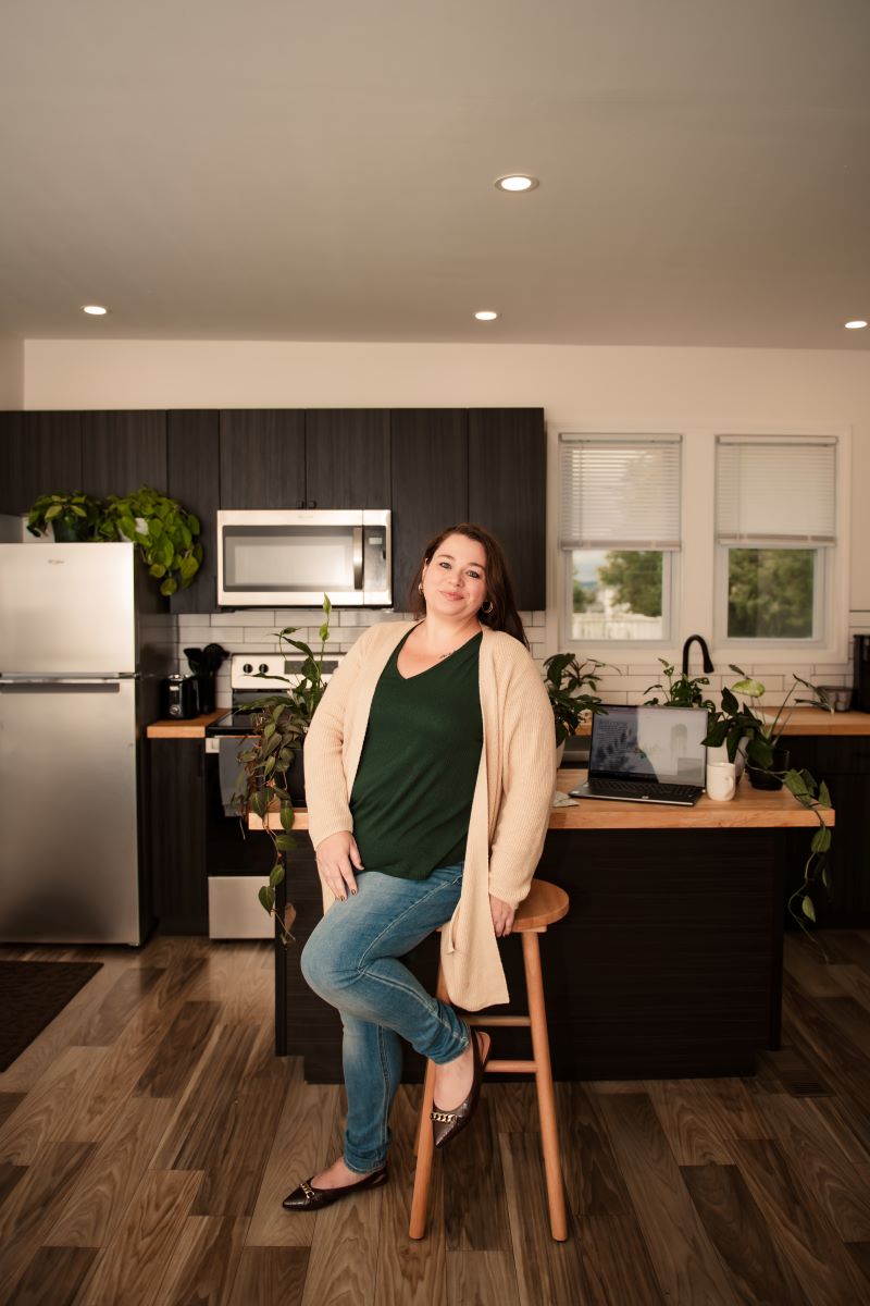 Emerald & Ivy owner, Chanell, leans on a wooden stool in front of a kitchen island. On the island are several plants, a coffee mug, and laptop that is opened up to Emerald-And-Ivy.com.